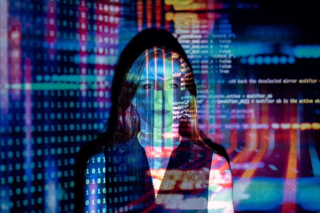 A woman faces the camera as brightly colored projections of computer code and mathematical notations are projected across her face.
