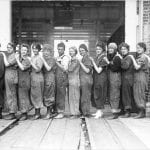 black and white photograph of a dozen women in worksuits, standing in a row with their hands on one another's shoulders
