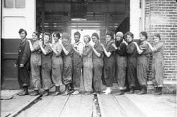 black and white photograph of a dozen women in worksuits, standing in a row with their hands on one another's shoulders